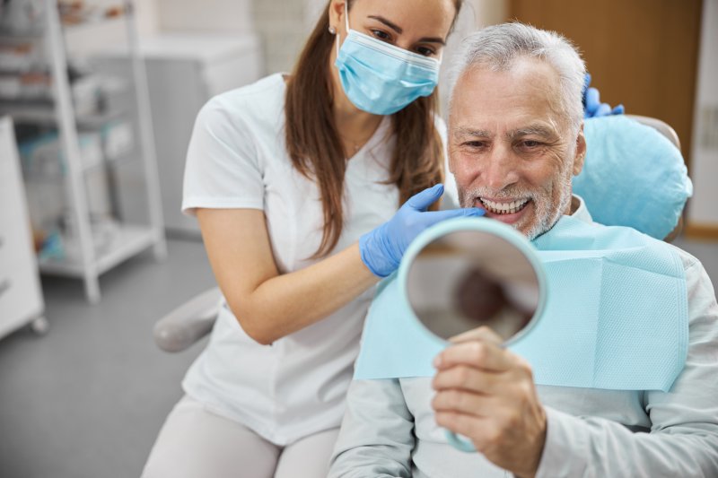 Dentist holding gloved hand next to an older man holding a mirror up to his smile